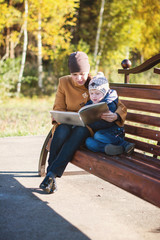 Young woman and little boy sitting on the bench in autumn Park and reading book. Family enjoying time together. Mother teaches son