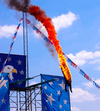 Fire Diver Daredevil Performing A High Dive Off Platform Head First Into A Pool Of Water. Stuntman Dressed In A Black Fire Protecting Outfit. A Dangerous, Extreme Yet Exciting Athletic Sport Stunt.