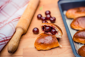 Oven fresh home made baked patties stuffed with cherry. Delicious pastries on a baking sheet, surrounded by cherry, napkin and rolling pin.