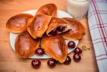 Oven fresh home made baked patties stuffed with cherry. Glass of milk. Delicious pastries on a baking sheet, surrounded by cherry, napkin and rolling pin.