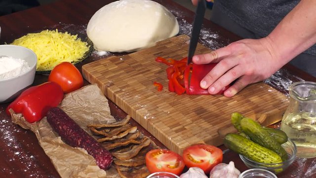 Mens hands cut red bell pepper on bamboo cutting board for cooking pizza. Pizza ingredients on wooden background