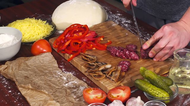Mens hands cut salami on bamboo cutting board for cooking pizza. Pizza ingredients on wooden background