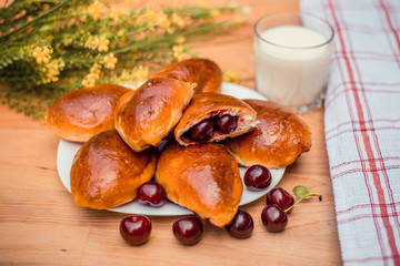 Oven fresh home made baked patties stuffed with cherry. Glass of milk. Delicious pastries on a baking sheet, surrounded by cherry, napkin and rolling pin.
