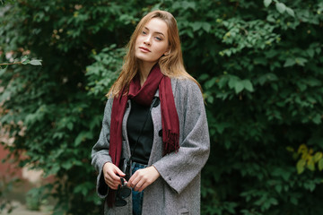 Beautiful stylish young woman in a warm scarf blue jeans trendy cozy coat posing alone against green foliage in fall city outfit. Touching her hair. Retro film filter edit.