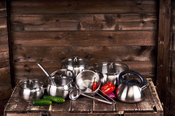 Different stainless steel pots, pans and laddles and fresh vegetables on old grunge wooden table against wood wall background