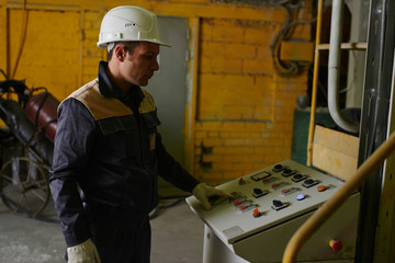 Male worker in overalls and helmet presses a button on the control panel at the premises of the plant