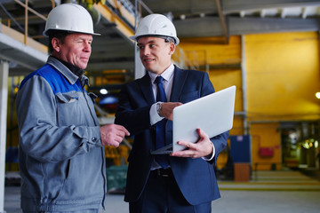 The man in the suit and the helmet holds the portable computer and shows up on the screen to the worker in overalls in an industrial building