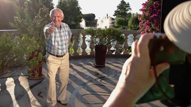Senior man shows thumb up. Woman's hand holding camera. Take a photo near fountain. Grandpa and grandma at resort.
