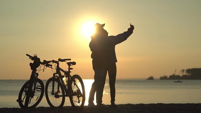 Silhouettes of two cyclists at sunset. The pair finished the bike ride on the river and do selfi on a beautiful background. Couple in love. Unusual date. hotograph on a mobile phone