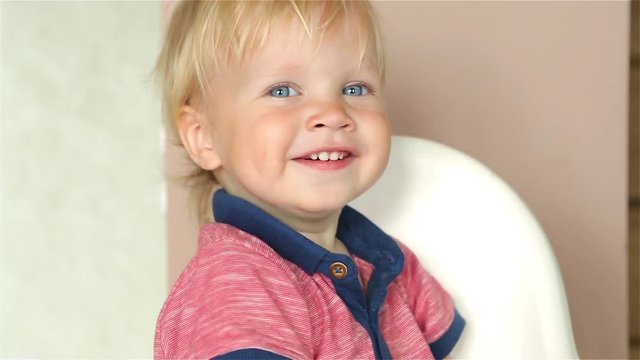 Adorable little happy baby boy is looking into the camera. The baby looks around and then smiles and laughs. 1 year old fair-haired child with blue eyes smiles at home, with sound (soundtrack)