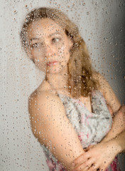 sexy young woman, posing behind transparent glass covered by water drops. melancholy and sad female portrait