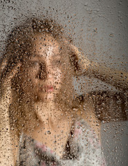 Fototapeta premium sexy young woman, posing behind transparent glass covered by water drops. melancholy and sad female portrait