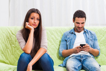 Young couple is sitting on sofa at home. Woman is getting bored while man is typing on phone. 