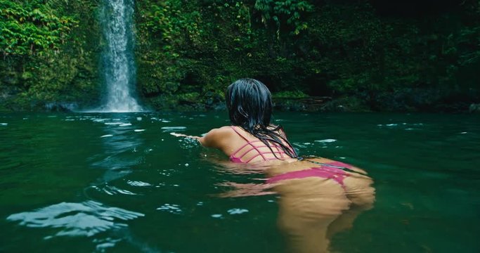 Young woman swimming under jungle waterfall