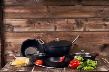 Different sblack pots, pans and stainless steel laddles and fresh vegetables on old grunge wooden table against wood wall background