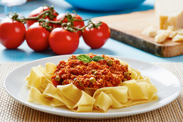 Tagliatelle with Bolognese sauce in white dish surrounded by fresh tomatoes and cheese on blue table