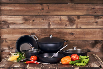Different sblack pots, pans and stainless steel laddles and fresh vegetables on old grunge wooden table against wood wall background