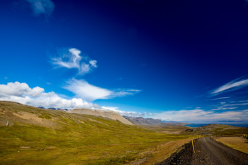 White clouds in the blue sky of West Iceland and a black gravel road with yellow poles marking the road