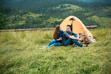 Fototapeta na wymiar A couple of tourists in time of the hike, sit outside the tent, mountain scenery. The guy hugs the girl. The concept of love and recreation
