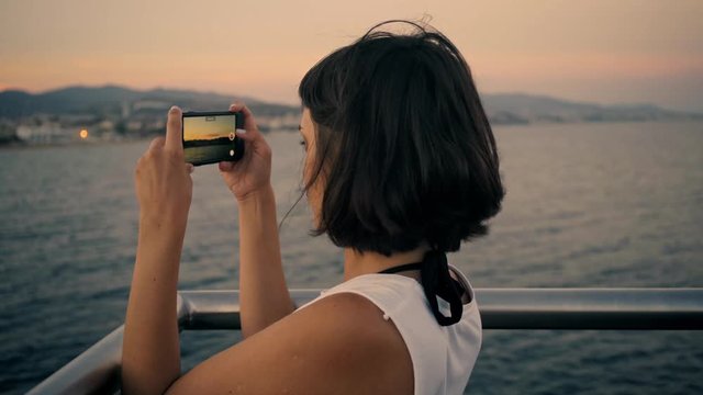 Beautiful brunette girl takes photo or video of sunset on her smartphone while standing on sea pier in front of city beach Wind sways her hair repeatedly Looped cinemagraph.