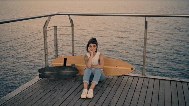 Pretty young brunette girl with tattoo on hand sits on wooden pier in front of longboard and small surf skate,looking at camera Wind sways her white t-shirt Beautiful sunset light Looped cinemagraph