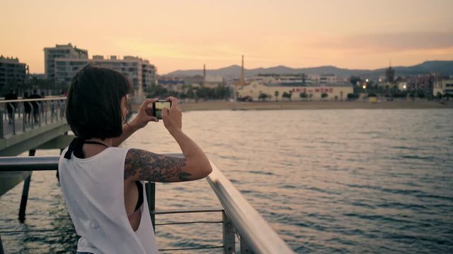 Attractive brunette girl with tattoo takes panorama view of sunset on her smartphone while standing on sea pier in front of city beach