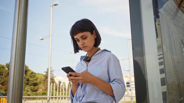 Beautiful brunette skinny girl in stripe oversived shirt waits her bus on public transport stop station in summer time and checking arrival schedule on her smartphone.