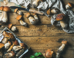 Freshly picked uncooked white forest mushrooms, green leaves and knife on rustic wooden background, top view, copy space, horizontal composition