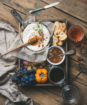 Cheese, fruit and wine set. Camembert in small pan with honey, nuts and herbs, grapes, persimmon, fig jam, baguette slices and glass of rose wine over rustic wooden background, top view