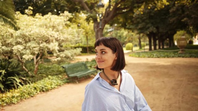 Happy smiling attractive brunette walks in park, looking at camera and sits on wooden park bench