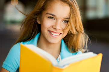 Smiling teenage schoolgirl reading book on highschool campus with wind blowing her hair