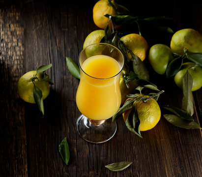 Glass of fresh tangerine juice with ripe tangerines, leaves and old-fashioned straw on a wooden background