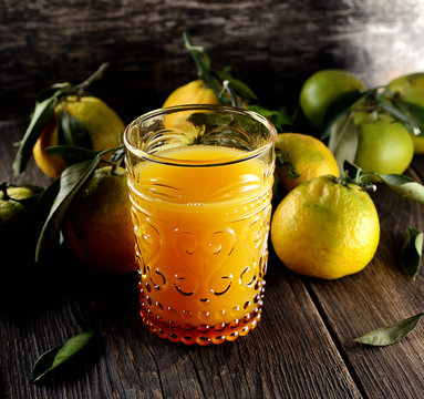 Glass of fresh tangerine juice with ripe tangerines, leaves and old-fashioned straw on a wooden background