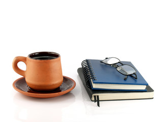 close up brown orange pottery coffee cup with saucer and two closed diary books (blue and black) with eyeglasses isolated on white background, read and write with hot drinks in baked clay cup