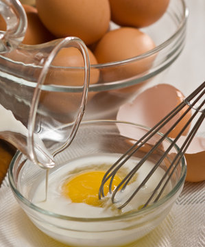 scrambling an egg and poring milk into a clear glass bowl with a whisk with cracked shell and whole brown eggs in glass bowl