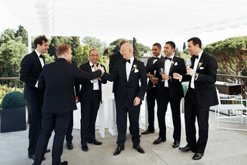 Bald-headed groom drinks wine with his friends under white pavil