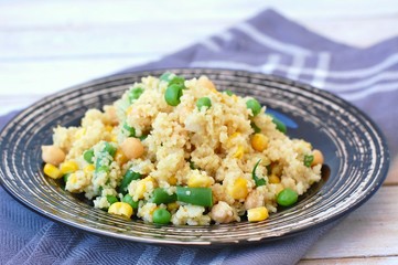 Healthy vegan meal with whole grain couscous, chickpeas, sweet corn, peas, green beans on dark plate on grey cloth on wooden background