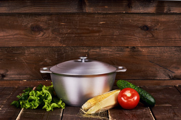 Cast aluminium pot standing on old grunge brown wooden table top. Lettuce, parsley, cucumber, tomato and corn