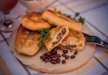 Fried tasty patties filled boiled beans and fried onion, on wooden plate, checked tablecloth on the background. Delicious buns on a baking sheet, surrounded by white sauce and glass of tomato juice.