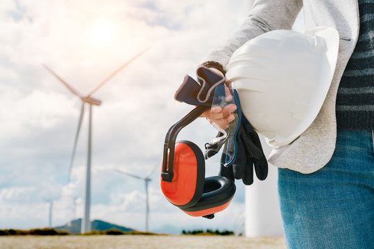 Engineer Woman Holding Safety Helmet, Gloves, Glasses And Ear Muffs At Wind Turbine Farm Generating Energy With Air Flow Spinning Blades. At Work Safety First Concept. Green Clean Energy.