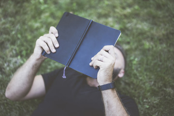 Summer day, a guy dressed in a black T-shirt lying on his back on the lawn in front of him holding a book in the black cover. Man lying on the grass and reading a book, his face hidden behind a book.