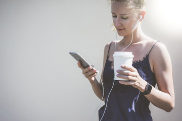 Sunny day,young woman dressed in blue top stands and using smartphone,while listening to music in headphones and holding cup with drink.On hand smartwatch,in background white wall.Girl using gadget.