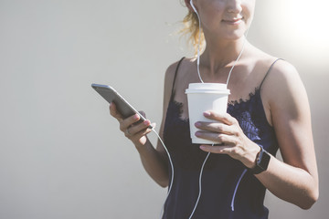 Summer sunny day,young smiling woman with blonde hair standing and listening to music on headphones,in hands holds smartphone and cup with drink,on the wrist smartwatch.In the background a white wall.