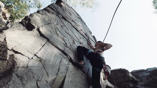 View from below of topless fearless climber ascending cliff with rope. He using carbines and long climbing rope