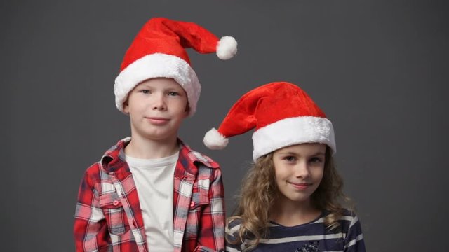 Couple of children in christmas caps looking to the camera and smiling, gray background