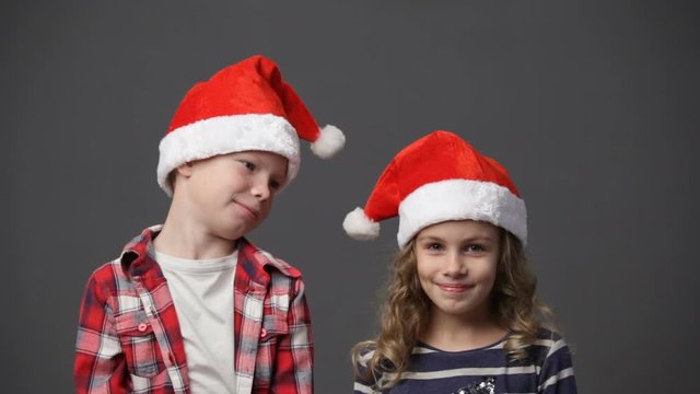 Little girl and boy in christmas caps looking at each other and then to the camera and smiling, gray background