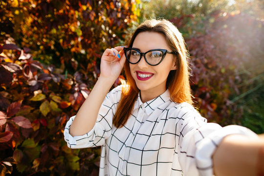 Gougeus model takes selfie while holding her glasses with one hand in autumn garden.