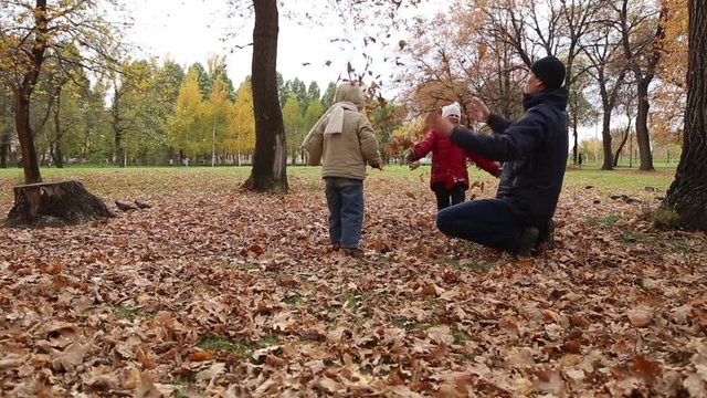 Father playing with daughter and son in autumn park on the fallen leaves, they are sit on the leaves and throw leaves at each other, daughter hugging father and they fall on the leaves

