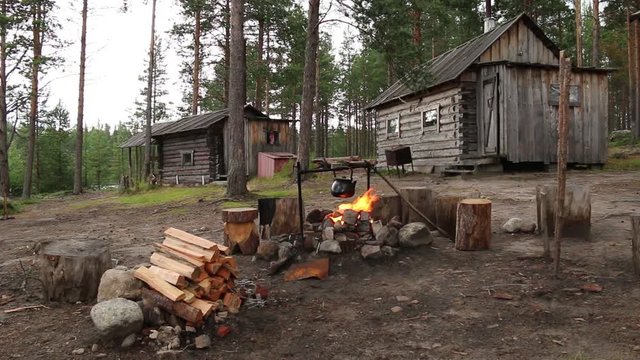 Kettle over the campfire on a background of the hut and a banya in the forest
