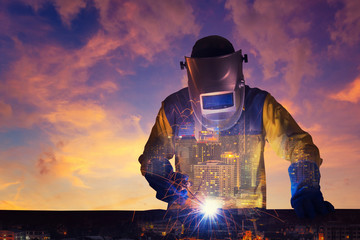 Double exposure of a downtown city and Industrial Worker welding steel structure with sunset sky in...